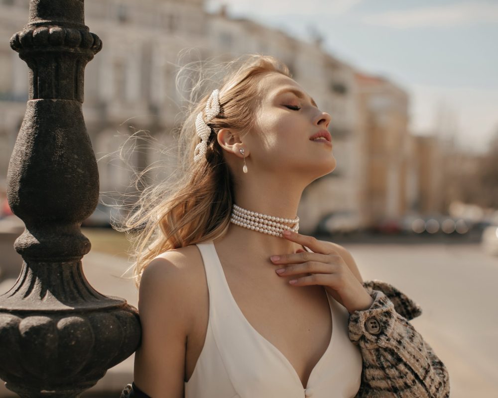 Portrait of beautiful blonde lady in stylish white silk dress, checkered jacket and pearl necklace gently touching neck and posing at city square.